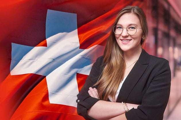 Porträt von Lara Wienbrauck, VAT Specialist bei exporto, vor einer großen Schweizer Flagge. Sie trägt eine Brille und einen schwarzen Blazer, lächelt in die Kamera und steht mit verschränkten Armen da. Das Bild betont ihre Expertise in Umsatzsteuer und grenzüberschreitender Compliance im Schweizer Markt.