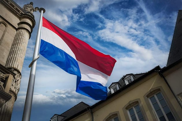 Nationalflagge der Niederlande in Rot, Weiß und Blau vor historischen Gebäuden und blauem Himmel – Symbol für Niederlande, Amsterdam, Europa und niederländische Architektur.