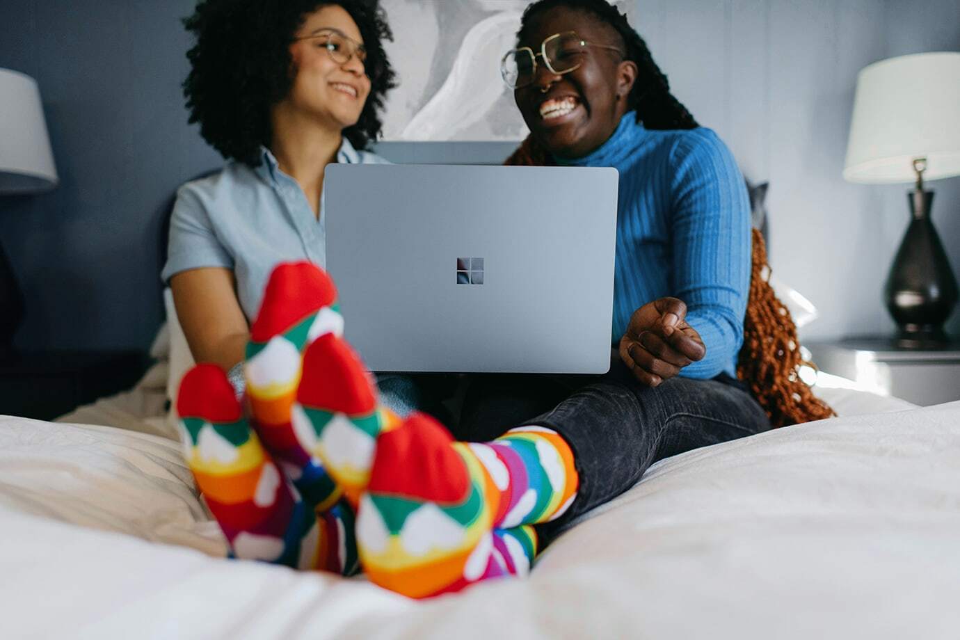 Zwei Frauen shoppen online am Laptop auf dem Bett in gemütlicher Atmosphäre, bunte Socken im Vordergrund – entspanntes Online-Shopping zu Hause.